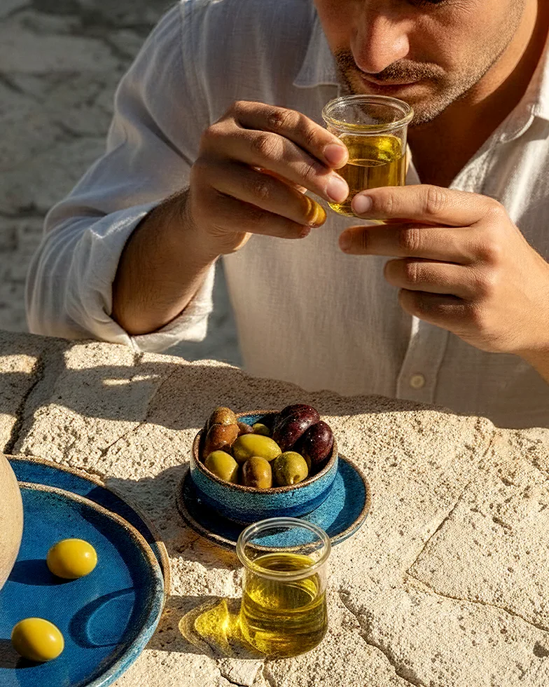 in einem mediterranen Setting riecht ein Mann an einem Glas mit Olivenöl, vor ihm steht ein Kurg mit frischem Öl und eine Schale mit frischen Oliven