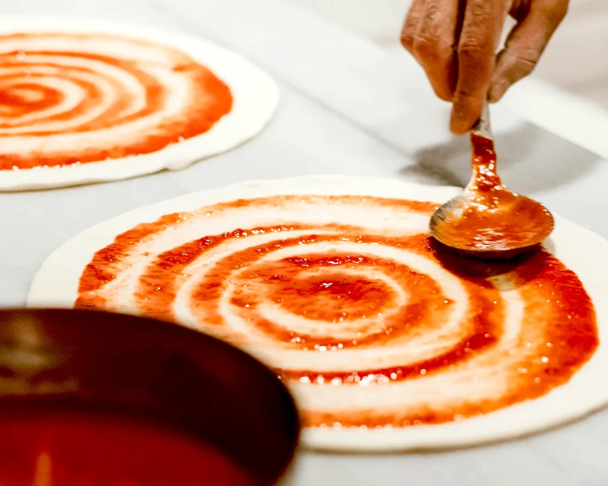 auf einer weißen Arbeitsfläche werden zwei Pizzateige in kreisförmigen Bewegungen mit Soße bestrichen