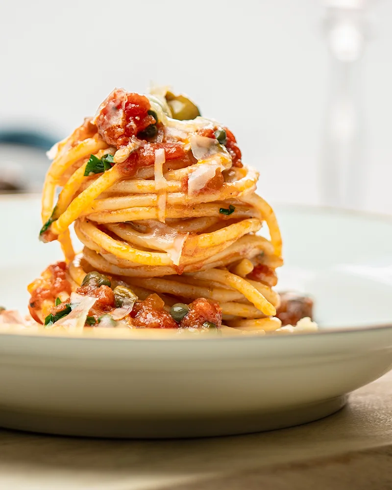 Spaghetti alla Puttanesca werden in der Seitenansicht auf einem weißen Teller serviert gezeigt