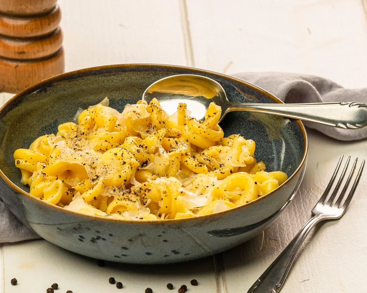 pasta cacio e pepe serviert auf einem blauen Steinzeug-Teller, dieser steht auf einem weißen Tisch eine Pfeffermühle steht im Hintergrund