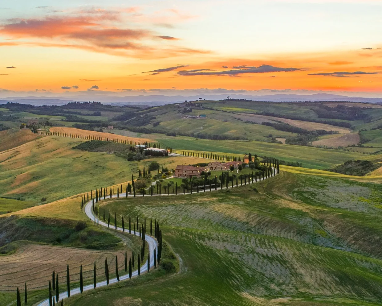 Sonnenuntergang im Panoramaformat: Das Valle D'Orcia mit grünen Hügeln, Zypressenbäumen und mediterranen Häusern liegt friedlich unter einem dramatischen Himmel in Orange-Blau-Violett