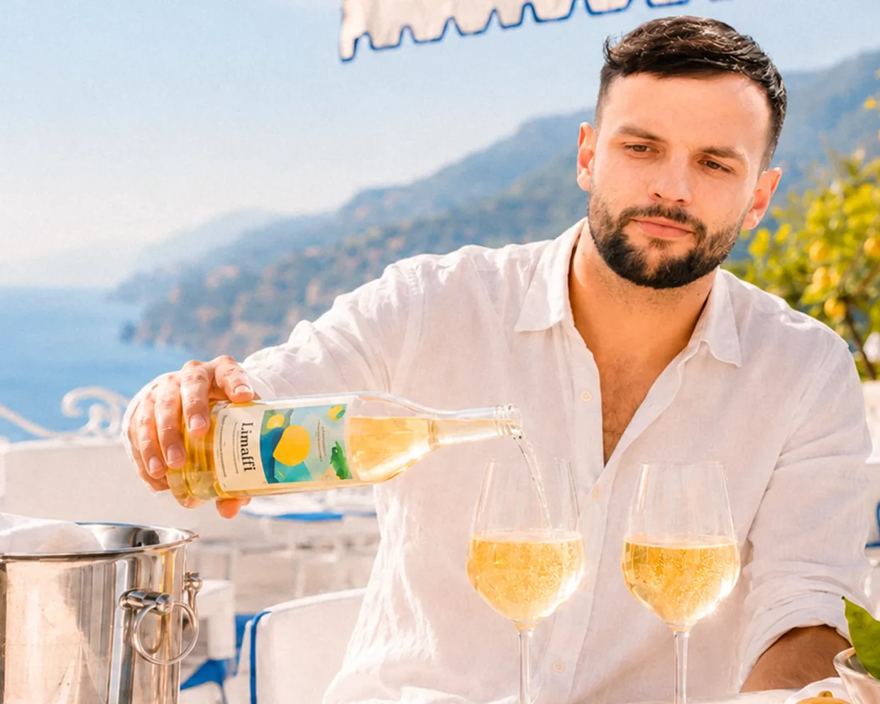 Franz schenkt auf einer sonnigen Restaurantterrasse an der Amalfiküste goldgelben Limalfi in zwei Gläser ein; daneben stehen Zitronen und ein Weinkühler.