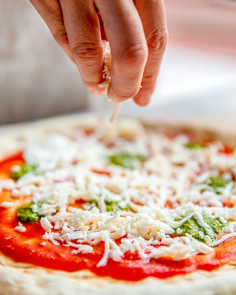 eine Hand streut Käse über eine Pizza
