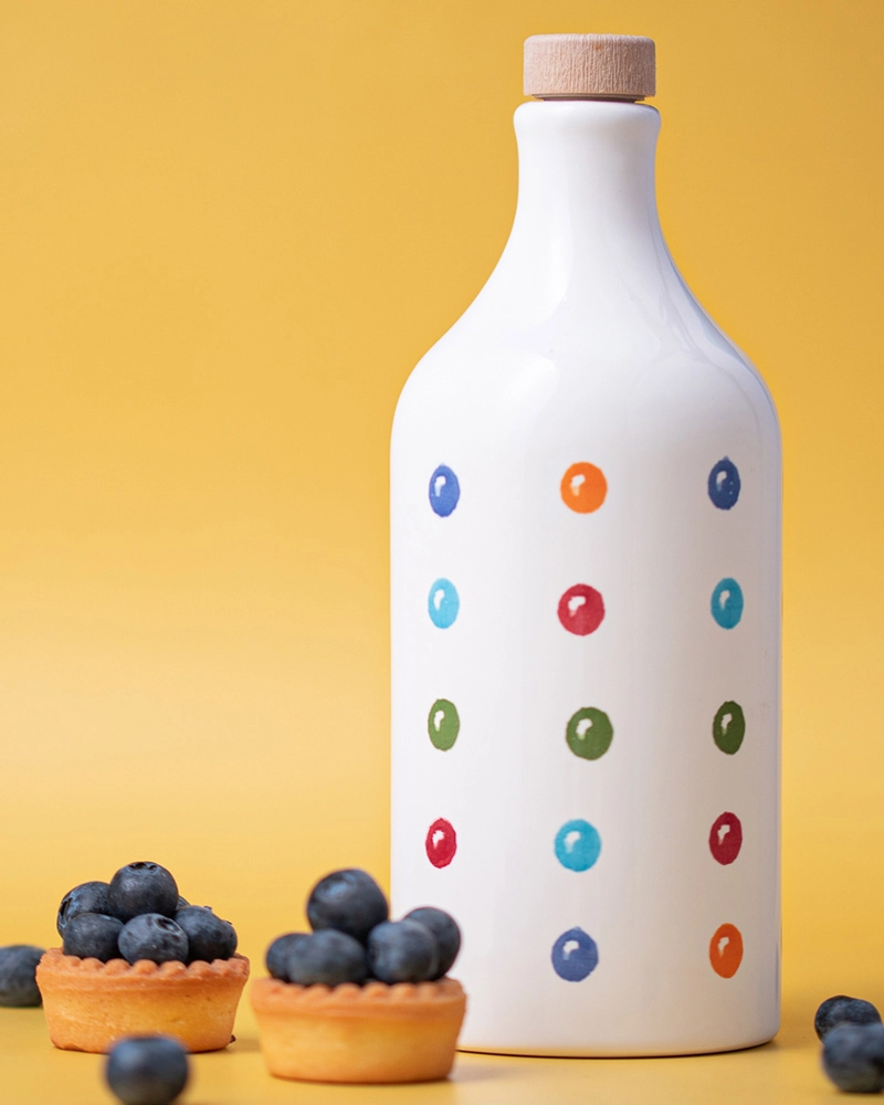 Muraglia Olivenöl - Punkte Keramikflasche Muraglia Olivenöl in gepunkteter Keramikflasche vor gelbem Hintergrund, Blaubeer-Törtchen im Vordergrund