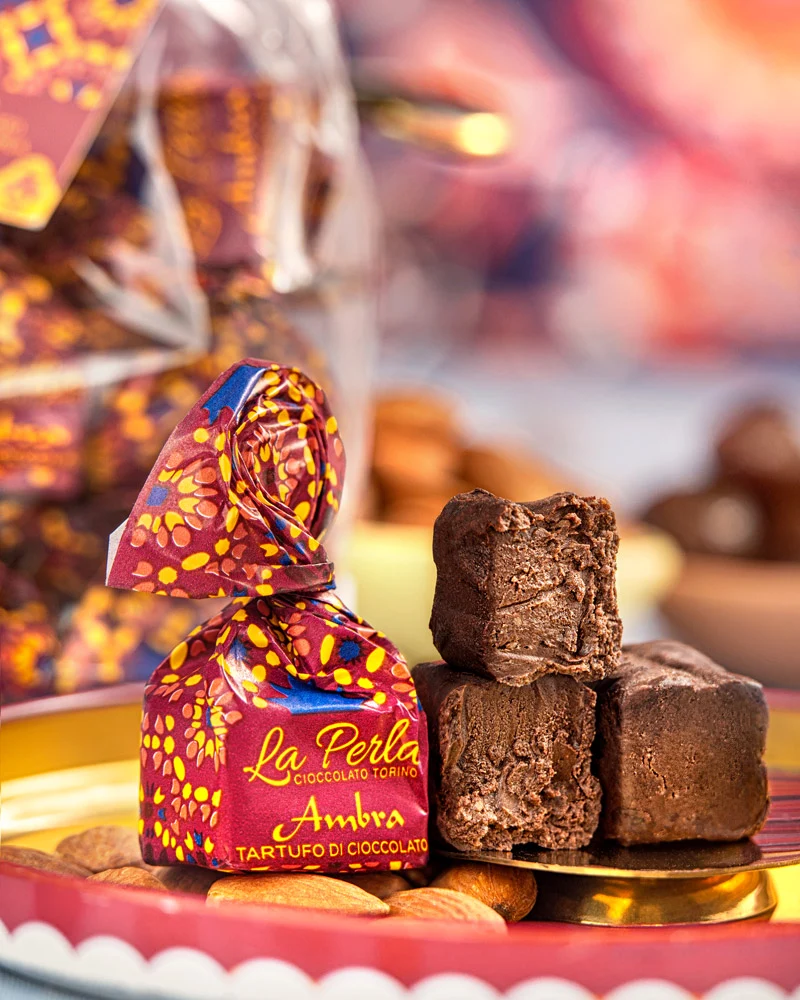 Tartufi Ambra - ohne Zuckerzusatz Ambra Trüffelpraline im Ornament-Papier mit drei geöffneten Pralinen auf goldenem Teller